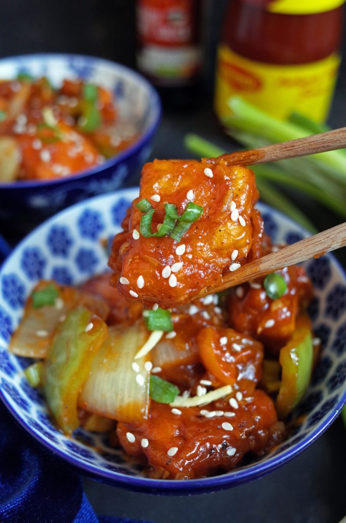 Close up photo of Indo-Chinese Chilli Tofu in a blue bowl. One piece is held by chopsticks and topped with sesame seeds