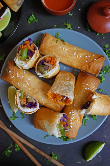 A plate of crispy vegan vegetable spring rolls with sesame seeds, lime and coriander. Spring rolls are cut open to reveal filling inside