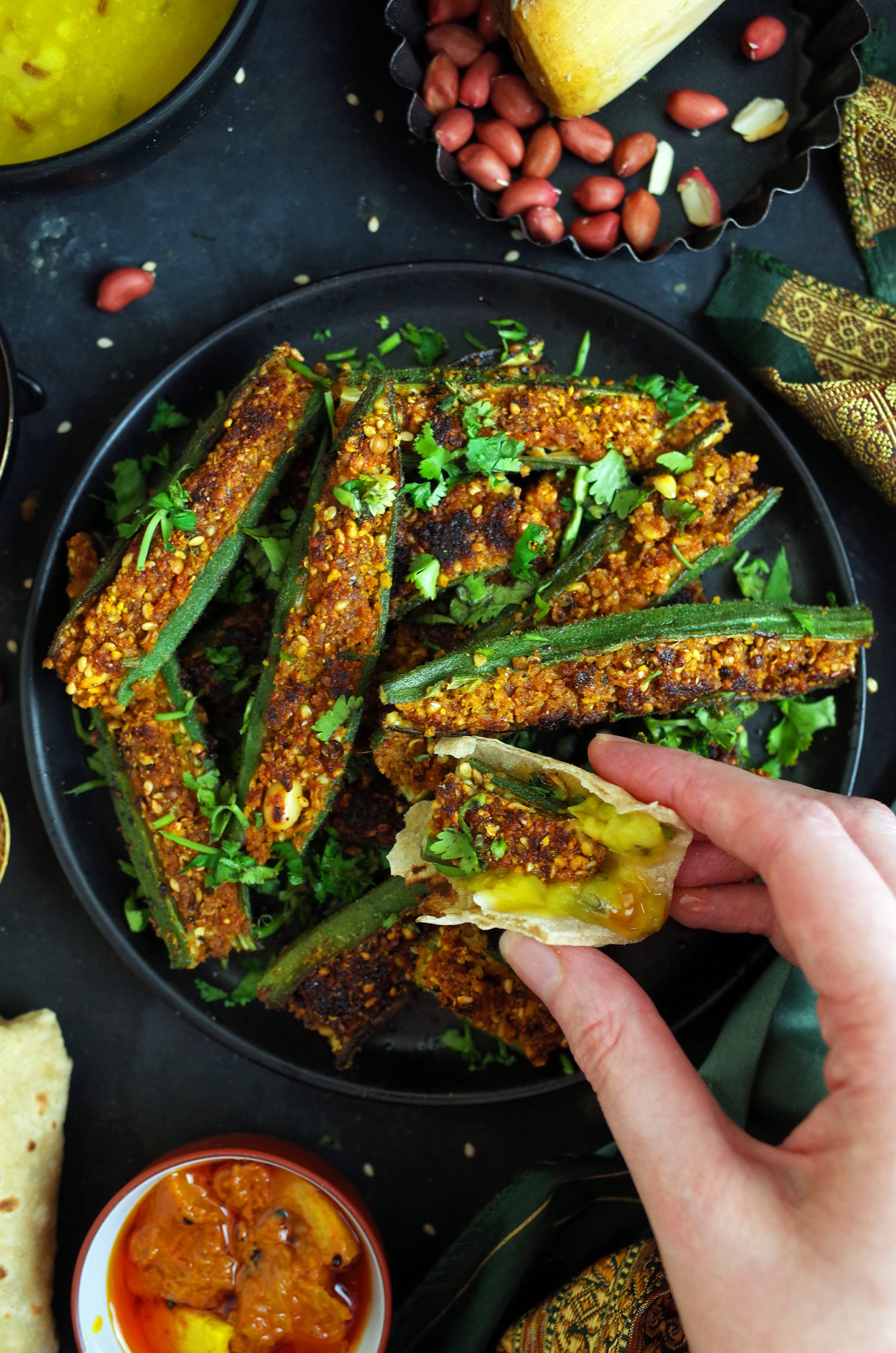 Maharashtrian bharli bhendi on a black plate. A hand is holding a piece of chapati with stuffed okra inside.