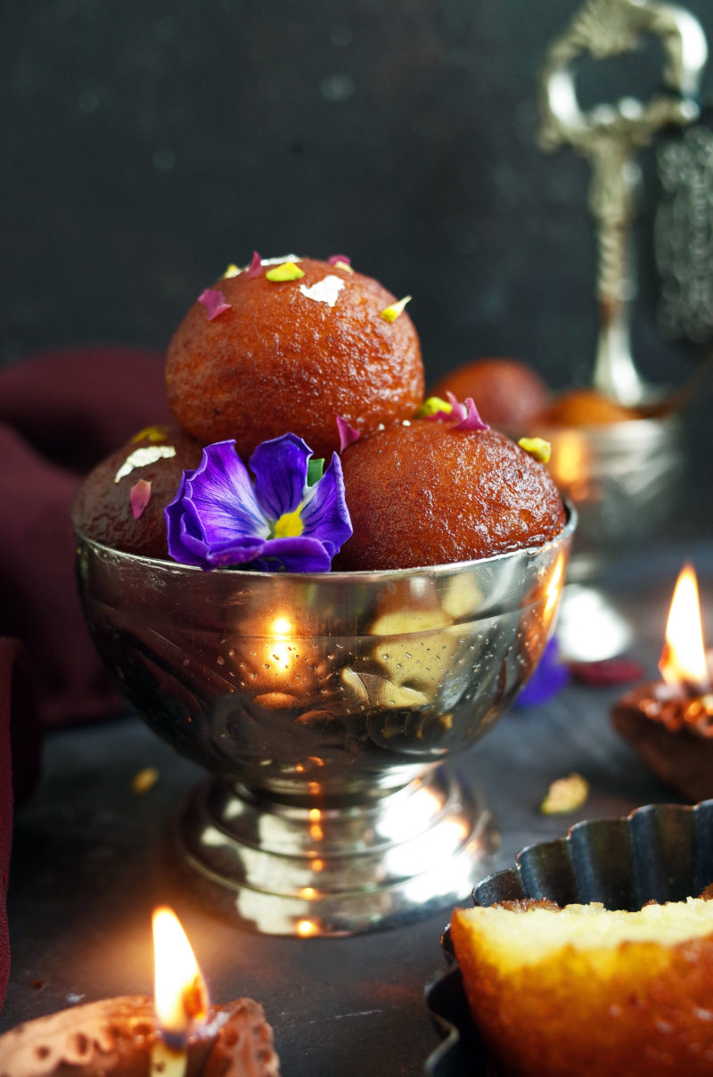 A pile of Indian Gulab Jamun topped with pistachios in a decorative metal bowl along with a pansy