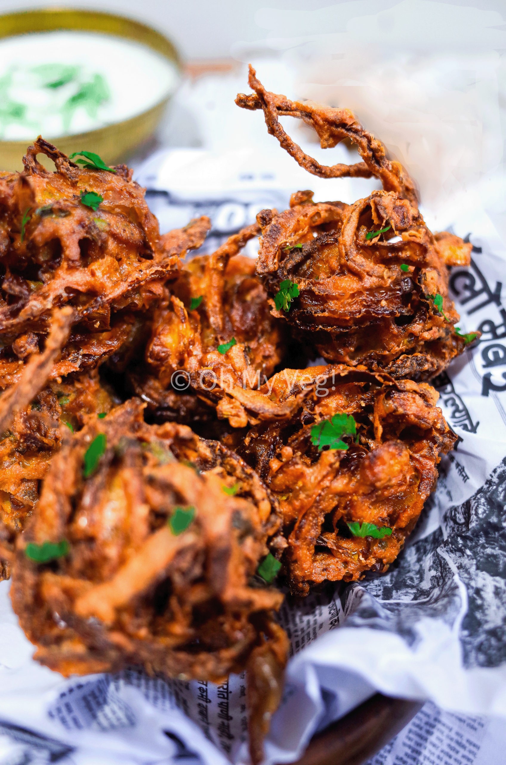 A pile of crispy onion bhaji with coriander on newspaper sheets with raita in the background