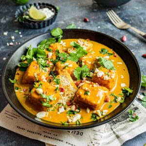 Tofu satay curry in a black bowl, with garnishes and a small bowl of pak choy in the background.