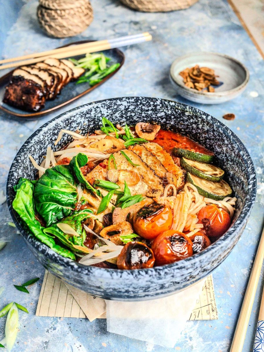 Vegan spicy ramen in a blue bowl, with toppings in the background. 