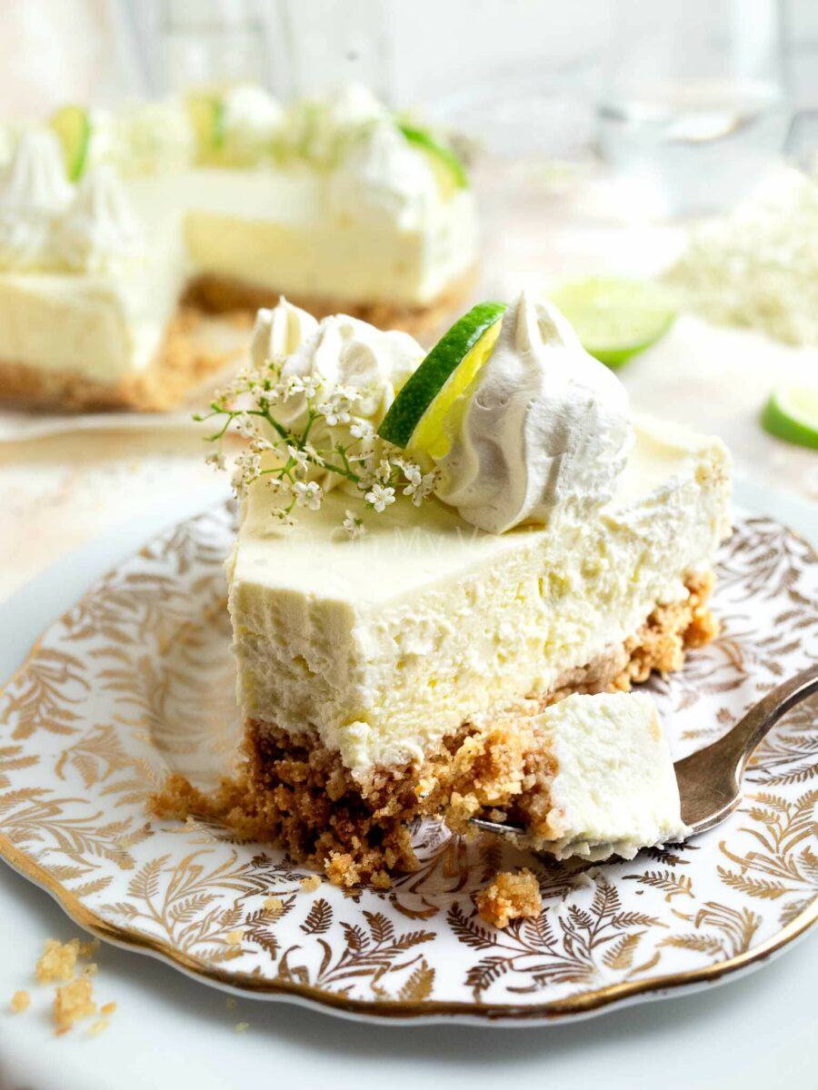 A piece of elderflower cheesecake with a fork, decorated with whipped cream and limes. 