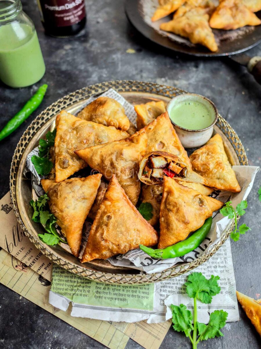 Chilli paneer samosas on a silver platter with chillies and sauce in a small pot. 